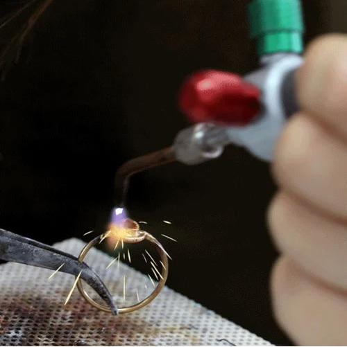 A close-up of a gold ring being held in tweezers while a jeweler uses a torch with a green cap to heat it, with sparks flying in a dark workshop setting.