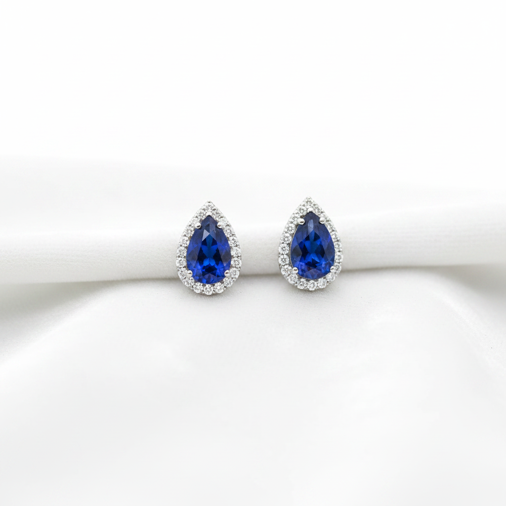 Pear cut blue sapphire halo stud earrings on white velvet, studio lighting